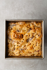 overhead view of onion and scallion focaccia in a baking tin, top view of airy bubbly onion focaccia on a cement countertop	