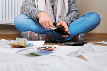 Man sitting on floor checking wallet near radiator surrounded by bills, euros and calculator due to high heating costs.