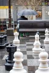 A man sitting in the rain with an unbrella and a giant chess set in the foreground, vertical