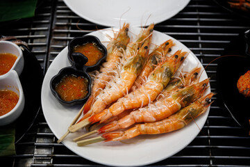Grilled shrimp skewers served on a white plate with spicy Thai seafood dipping sauce. Delicious street food delicacy featuring fresh, vibrant orange prawns ready to eat.