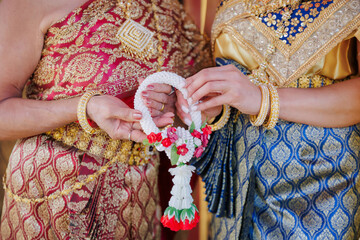 Two women in traditional Thai silk dresses holding a colorful handmade flower garland together. Elegant close-up of cultural heritage, jewelry, and friendship symbols.
