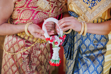 Two women in traditional Thai silk dresses holding a colorful handmade flower garland together. Elegant close-up of cultural heritage, jewelry, and friendship symbols.