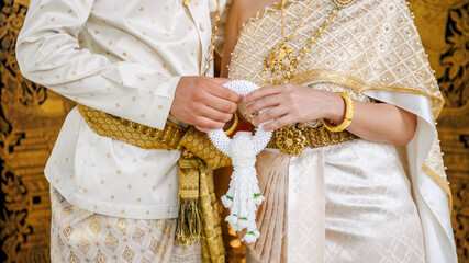 A Thai couple in traditional wedding attire holding a white jasmine garland together. Close-up of elegant gold embroidery, jewelry, and cultural marriage symbols.