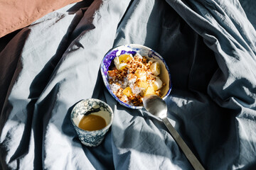 Yogurt bowl with fruit placed on rumpled bedding lit by sunlight