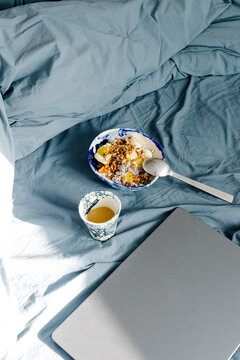 Fruit yogurt bowl placed on rumpled bedding next to a mug and a laptop