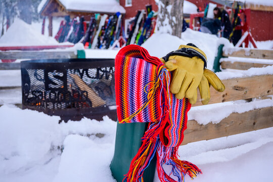 Colorful traditional Quebecois "ceinture fl&eacute;ch&eacute;e" arrow sash scarf and yellow leather work gloves resting on a snow-covered wooden bench by a fire pit at a winter ski resort.