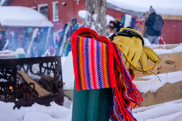 A colourful woven sash and a pair of yellow leather gloves rest on a wooden rail at an Ontario Nordic ski resort in winter, with ski gear and a fire pit visible in the snowy background.