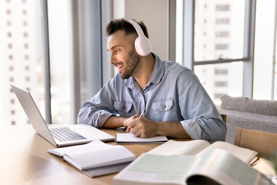 Young 30s Hispanic man listen to audio class using modern wireless headphones, taking notes in notebook, engaged in online course, participate in webinar for gaining new knowledge remotely. E-learning