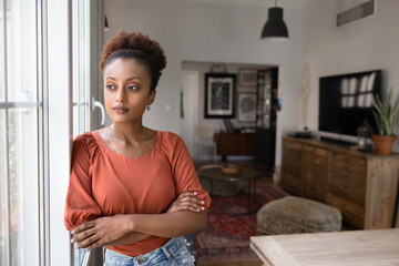 Serious thoughtful young African woman standing at window, keeping stressful close pose with hands...