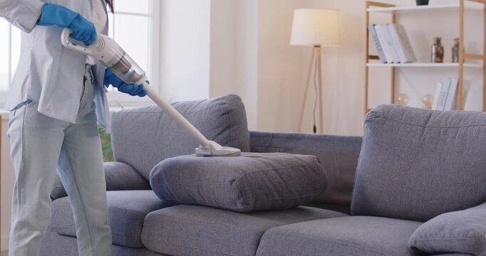 Woman vacuum cleaning sofa cushions during home cleanup. Hands of professional cleaner worker with gloves holding stick of cordless electrical appliance to remove dust in furniture, clean room - Powered by Adobe