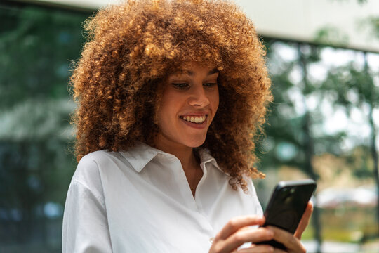 Smiling Woman With Curly Hair Reading Message On her Phone Outdoors