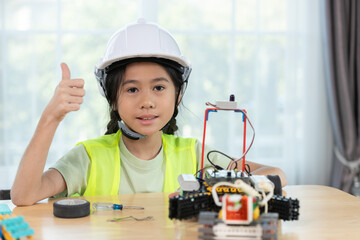 Confident girl in hard hat and safety vest giving thumbs up next to her completed robotic project, representing STEM success, innovation and encouragement for girls in tech.