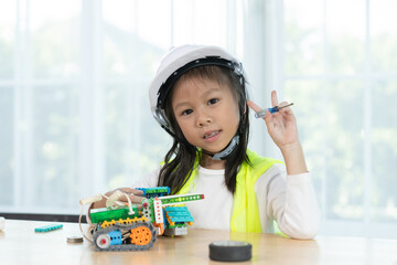 Happy girl in hard hat and safety vest holding tools and building colorful robotic machine, representing STEM learning, creativity and early engineering inspiration for kids.