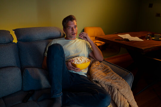 Man relaxing on sofa watching movie and eating popcorn at home
