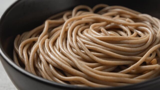 Cinematic focus rack of buckwheat soba noodles in a black bowl with wooden chopsticks. Close up of healthy Japanese pasta on a grey textured background.