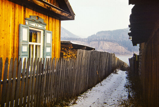 Wooden house and fence in snowy rural village, mountains and river 