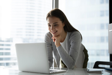 Inspired specialist at work. Smiling young female business professional executive stand leaning at office desk use notebook for videocall communication interact via text chat review online content