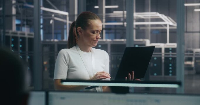 Mobile IT Management, Focused Female IT Professional Standing and Actively Working on Thin Laptop. Background Modern Server Hall. Emphasizes Mobile Enterprise Management and High Level IT Operations.