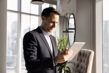 Positive young Hispanic executive man typing on tablet, standing in office, holding gadget, using online technology, financial application for managing business process, enjoying communication