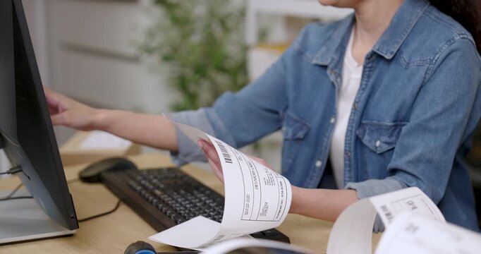 Asian female entrepreneur pulls long shipping labels from printer while checking customer orders on computer during e-commerce startup daily work inside home warehouse storage area