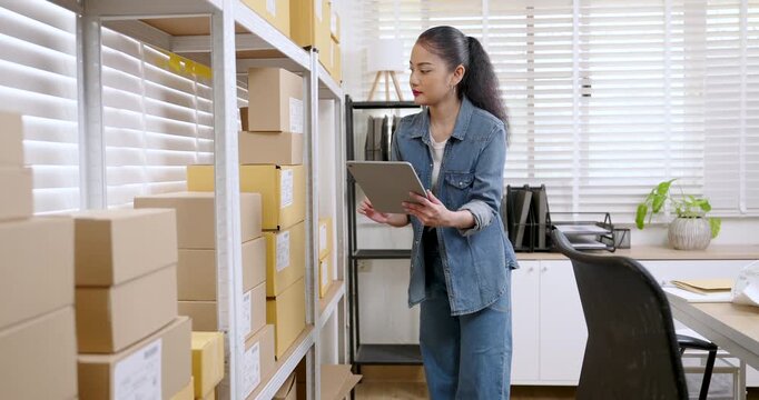 Asian woman small business owner moves down aisle, matches parcel boxes with customer order note, updates inventory spreadsheet, keeps e-commerce business workflow ready for packing products shipment