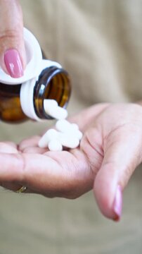 Vertical close up of a woman's hand holding a medicine bottle while taking vitamin pills and supplements like probiotics or magnesium to support her daily health, wellness, and medical wellbeing