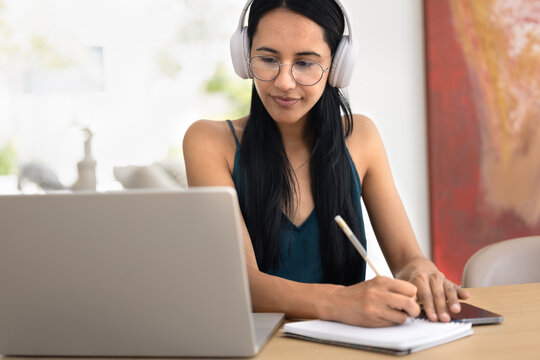 Focused young New Zealander female student in wireless headphones and eyeglasses watching online video lecture, learning webinar on laptop at home, writing notes in copybook