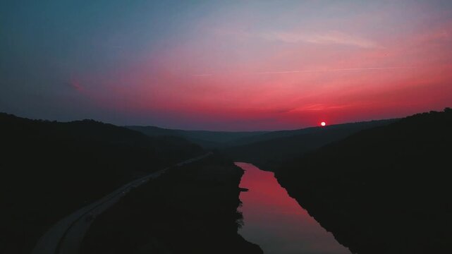 Cinematic slow downward shot of a small river at dusk, reddish sunset reflections on calm water, sun hiding behind hills, dark serene mood with a parallel highway adding subtle urban contrast.