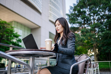 Japanese businesswoman working on laptop outdoors with coffee cup