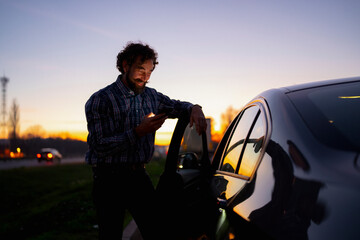 Man checks phone while leaning against car during sunset 