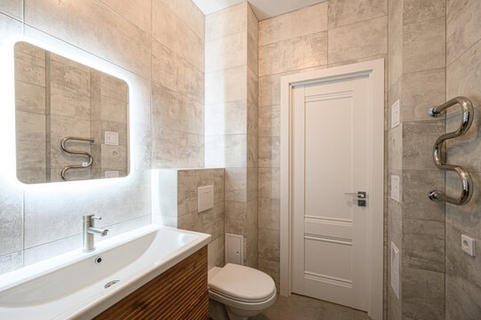 Modern bathroom with grey textured tiles, white sink with wooden vanity, illuminated mirror, wall-mounted toilet, chrome towel rail, white door, minimalist design - Powered by Adobe