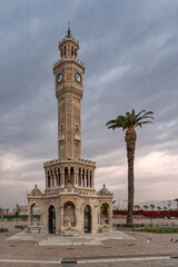 Obraz premium Iconic Izmir Clock Tower, a historic landmark, stands tall with a palm tree in Konak Square, Izmir, Turkey, under a dramatic cloudy sky. Pigeons gather on the ground.