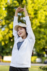Asian woman wearing wireless headphones, smiling while relaxing in a park after workout and stretching, enjoying music and peaceful outdoor moment of contentment and well-being.