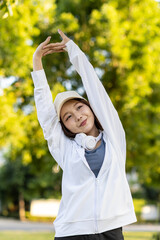Asian woman wearing wireless headphones, smiling while relaxing in a park after workout and stretching, enjoying music and peaceful outdoor moment of contentment and well-being.