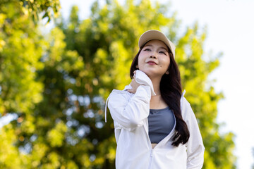 Asian woman wearing wireless headphones, smiling while relaxing in a park after workout and stretching, enjoying music and peaceful outdoor moment of contentment and well-being.