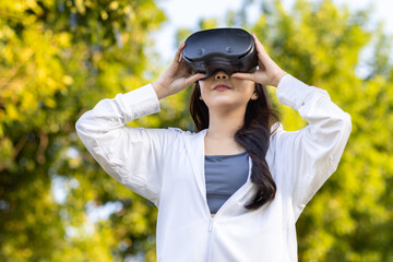 Asian woman wearing wireless headphones, smiling while relaxing in a park after workout and stretching, enjoying music and peaceful outdoor moment of contentment and well-being.
