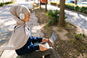 Young woman wearing hijab listening music working laptop outdoors