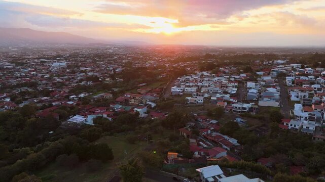 Aerial drone panning shot from right to left over the residential houses and lush green mountains of San Rafael de Heredia, Costa Rica, during a bright tropical sunset with a cloudy sky