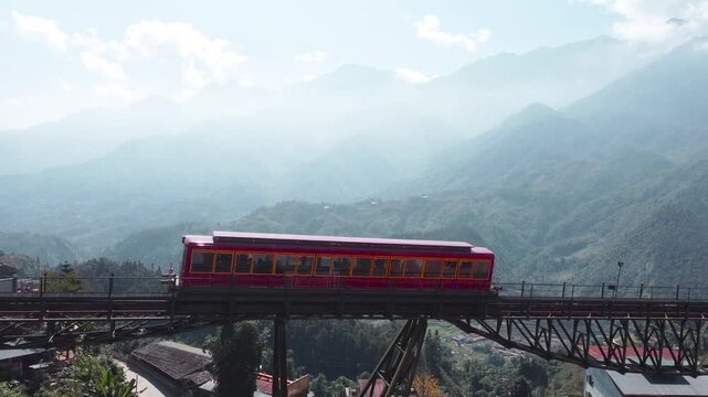 Red train travels on elevated bridge over green valley with misty mountains.