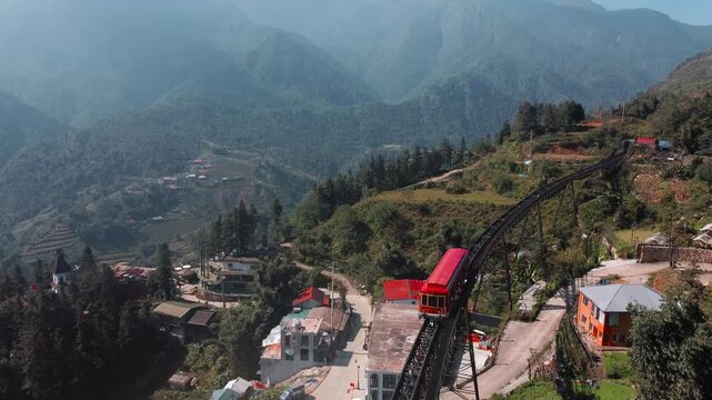 Red train travels on high track over village houses, misty mountains and green valley surround winding railway route.