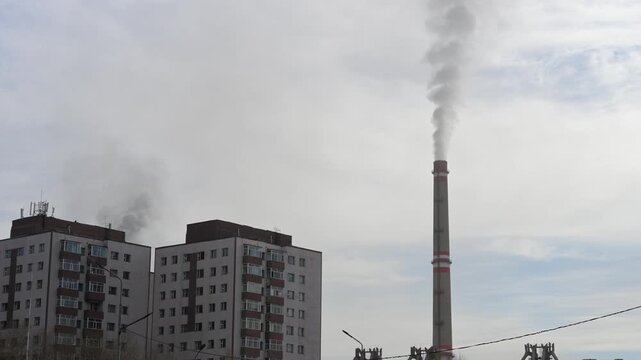 A tall industrial chimney emits smoke next to residential buildings in Ulaanbaatar, Mongolia. The power plant infrastructure looms over the city's living quarters and skyline.