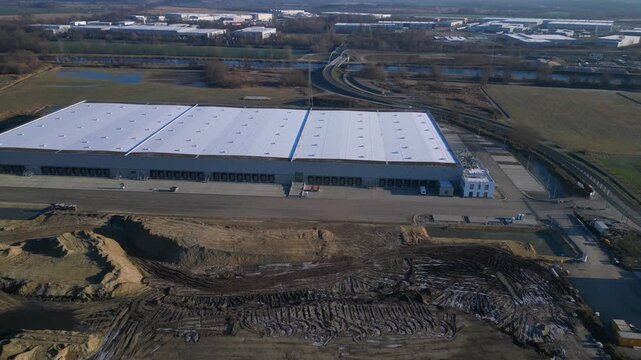 New large logistics center building under construction with a vast storage area, loading docks, and surrounding empty land. Gorgeous aerial view speed ramp hyper motion time lapse panorama orbit drone