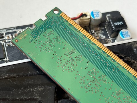 Close-up of a green RAM memory module with gold-plated contacts, placed diagonally on a computer motherboard. Clear view of circuit traces, chips, and labeling on the PCB.