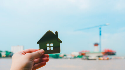 Real estate and property investment concept image with a hand holding house icon against blurred background of construction site and river, symbolizing housing market, home ownership.