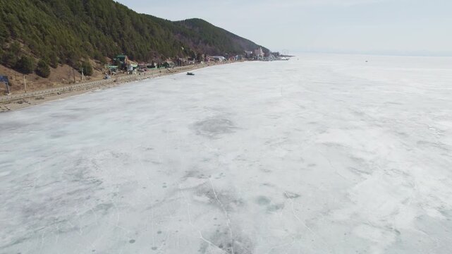 Flight over the ice of Lake Baikal in spring, the village of Listvyanka. Cars with tourists are driving along the road along the lake, and hovercraft are driving on the ice.