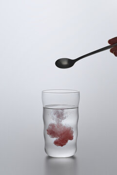 A spoon adding a red substance to a glass of water against gray