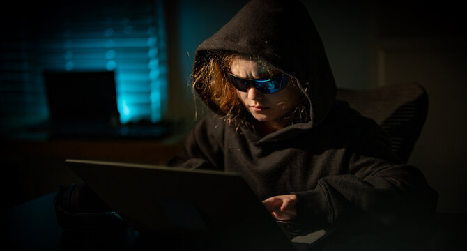 Young hacker with hood and glasses at night. Child hacker at night. Teen hacker working on computer in dark room. Kid hacker using laptop in a dark room. - Powered by Adobe
