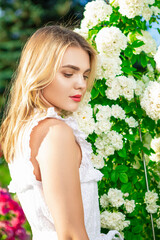Cute Airy Light Caucasian Fashion Blond Girl in Fragrant Pink Flowers Posing in White Dress in Summer Blossom Park as Fashion, Flowering, Cosmetics and Perfumes.