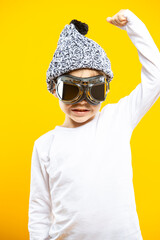 Positive Caucasian Boy Wearing Aviation Protective  Goggles and Warm Winter Knited Hat With White Shirt Lifted Hand As Strong Power Gesture Isolated On Yellow Background as Image To Portray Childhood