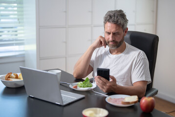 Businessman eats a morning meal while checking his laptop in a cozy home office. A mature business man planning his workday. A freelancer eats a breakfast. A casual man eating lunch at home office.